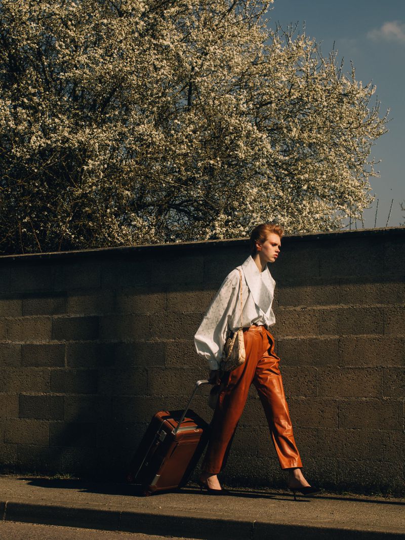 Fashionable redhead woman pulls a brown suitcase on a city sidewalk near a textured wall and flowering tree in bright daylight.