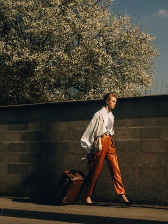 Fashionable redhead woman pulls a brown suitcase on a city sidewalk near a textured wall and flowering tree in bright daylight.