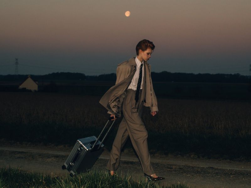 An adult in a trench coat and suit walks at dusk, pulling a rolling suitcase along a path in a rural setting, with a full moon in the sky.