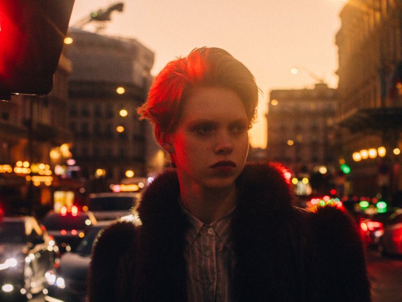 Portrait of a stylish young adult in a fur coat standing in a brightly lit city street at night with building and car lights.