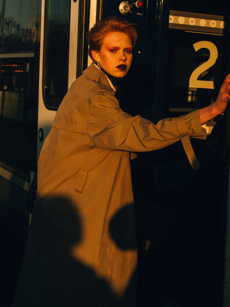 Stylish woman in a tan trench coat stands by a dark train door in a close-up fashion shot with a warm, moody lighting.