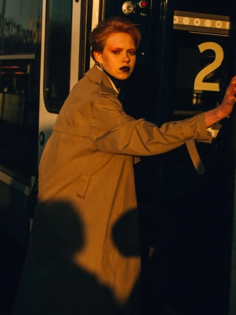 Stylish woman in a tan trench coat stands by a dark train door in a close-up fashion shot with a warm, moody lighting.