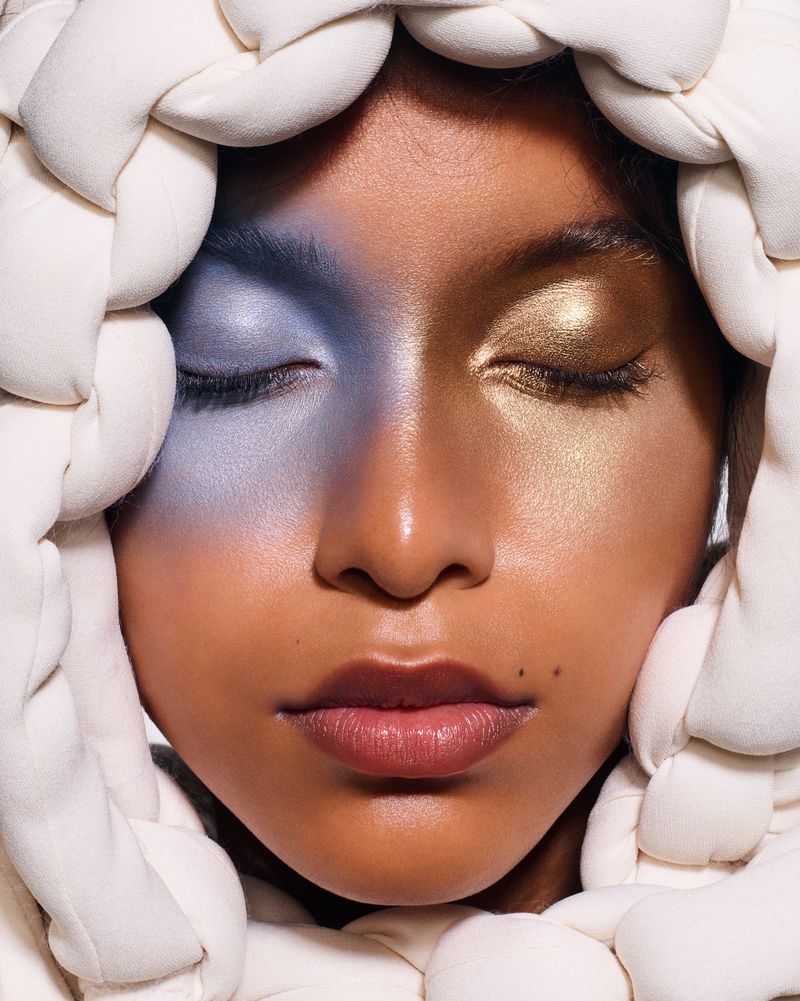 Close-up portrait of a dark-skinned woman with blue and gold eyeshadow, framed by a white, braided head covering, eyes closed.