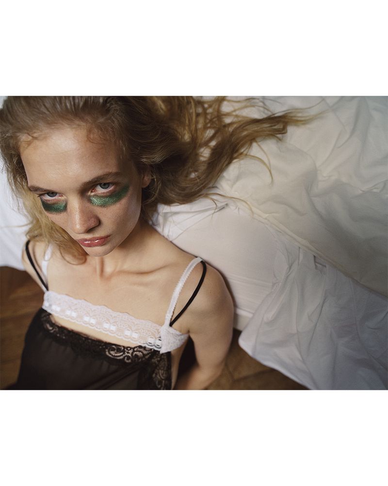 Model with green eye makeup wearing a black lace camisole reclining on white bedsheets.