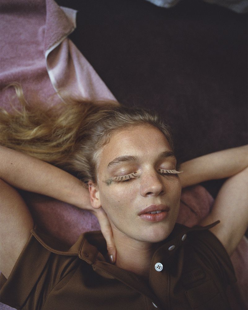 Close-up portrait of model Caitlin Soetendal with eyes closed, wearing a brown collared shirt against a pink towel and dark background.
