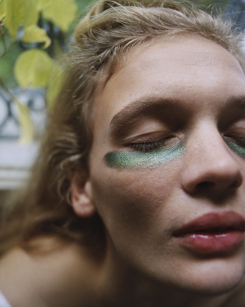 Close-up portrait of a woman with metallic green eyeshadow streaks beneath her eyes and closed eyes.