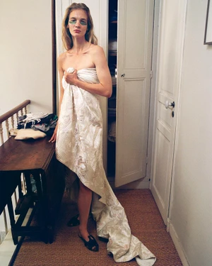 Model wrapped in a patterned white fabric sheet standing in a domestic hallway next to a dark wood console table.
