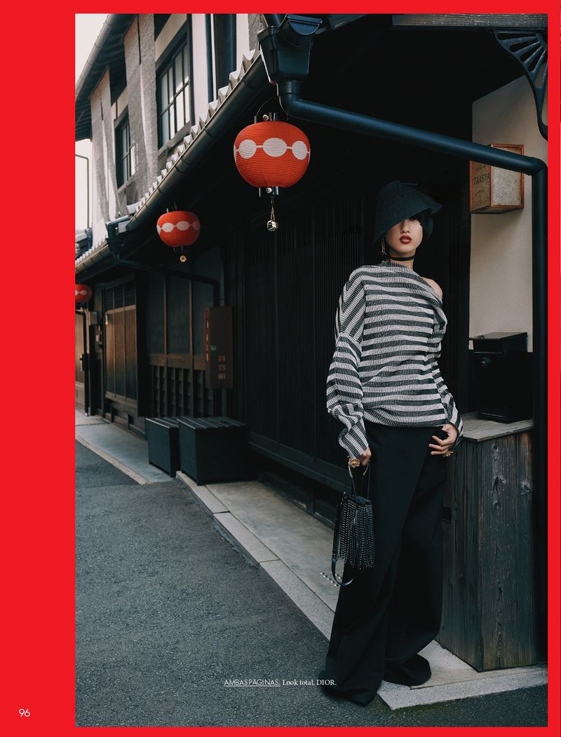 Fashion shot of a woman in a striped sweater and wide-leg pants poses outdoors against a dark building with red lanterns.