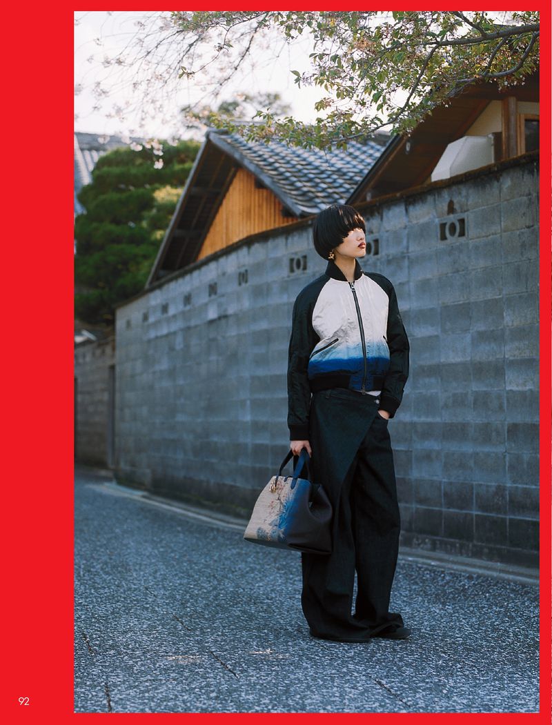 Adult female in bomber jacket, wide-leg pants and tote bag stands in a stone alleyway with traditional Japanese architecture.