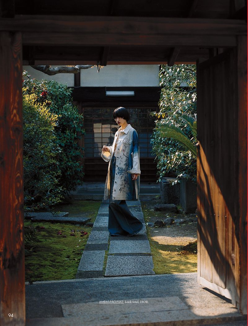 A fashionable person stands on a stone path in a verdant Japanese garden, wearing a long, patterned coat and wide-leg pants, framed by a wooden gate.