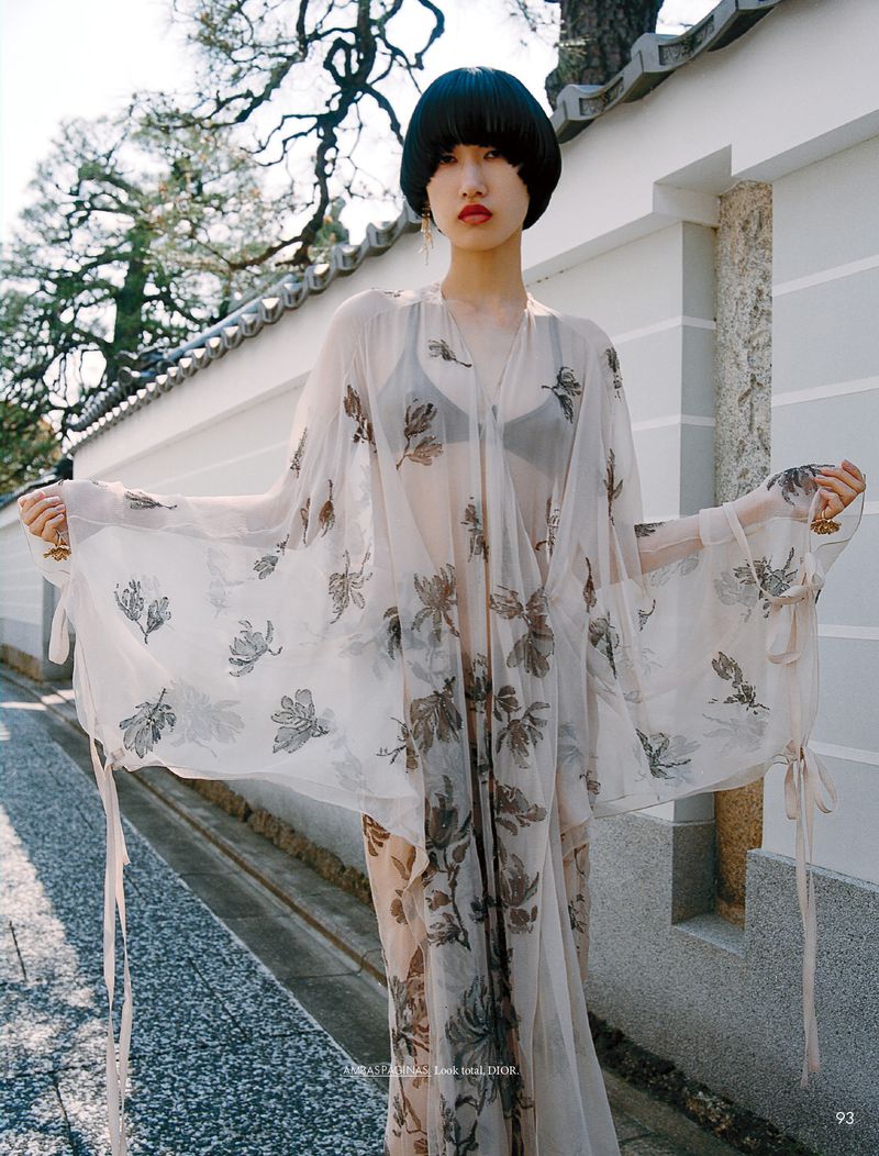 A young woman with dark hair poses in a patterned dress with flowing sleeves on a street with a wall and trees in the background.