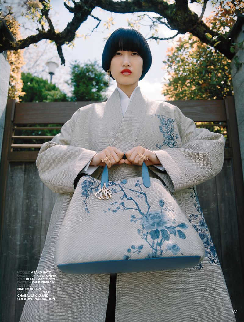 Adult female with black bowl-cut hair poses outdoors wearing a tan coat with blue floral pattern and holding a light-blue tote bag.