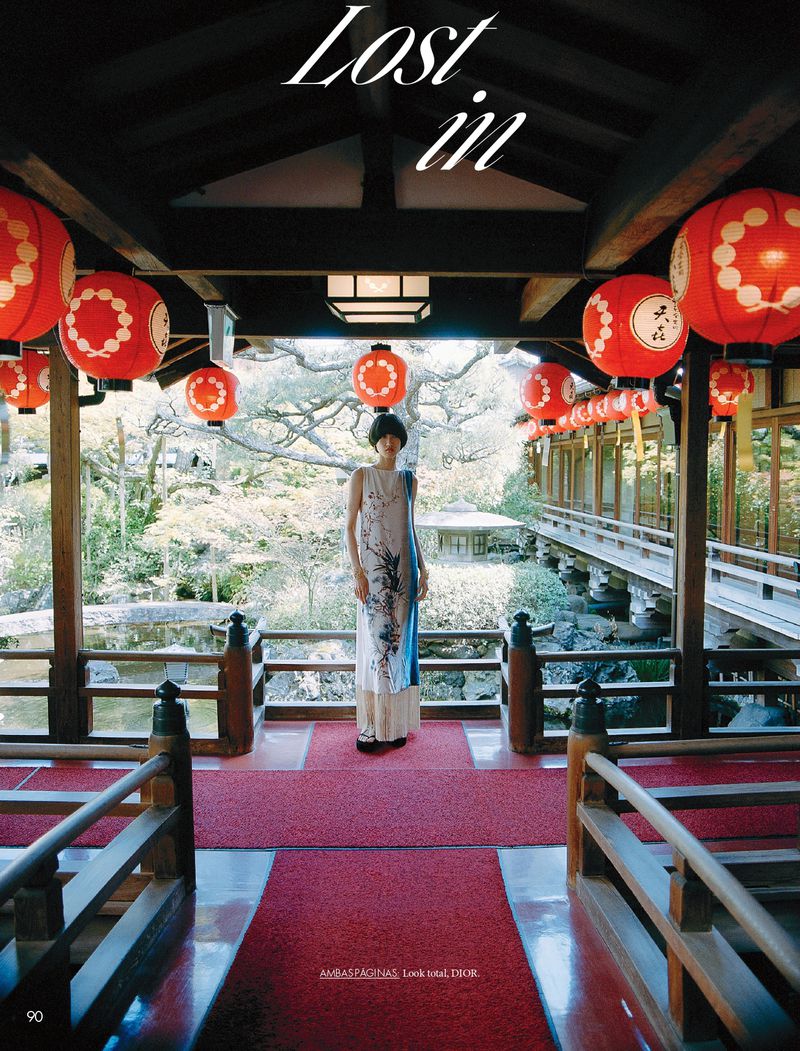 An Asian woman in a long dress stands inside a building that opens to a Japanese garden with red lanterns hanging from the ceiling.