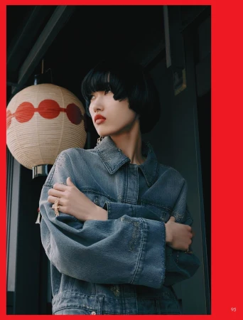 Asian woman in a denim jacket posing indoors near a paper lantern with red accents, styled with bold jewelry and dark blunt bob hairstyle.