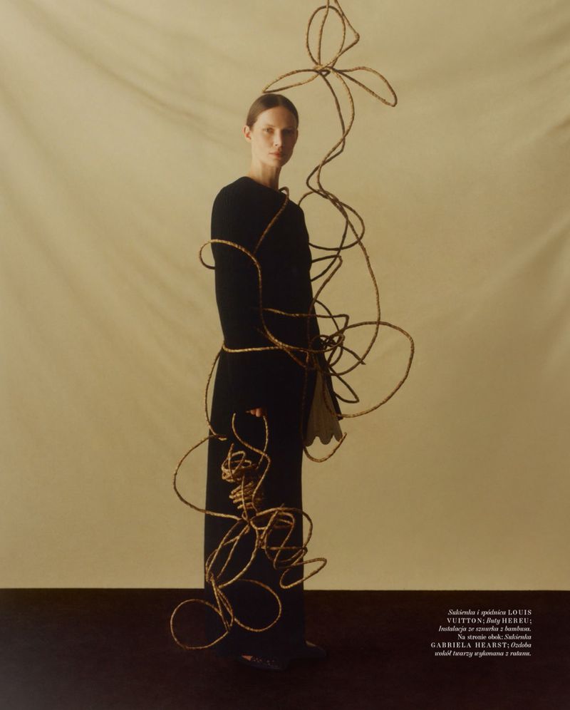 Model in a black dress standing against a neutral backdrop intertwined with looping wicker sculpture elements for Vogue Polska