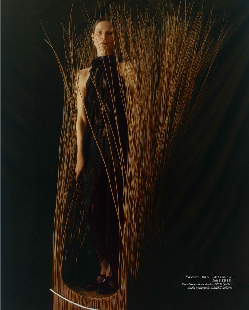 Model standing inside a sculptural vertical woven wicker structure against a dark background for Vogue Polska project Plecionka.