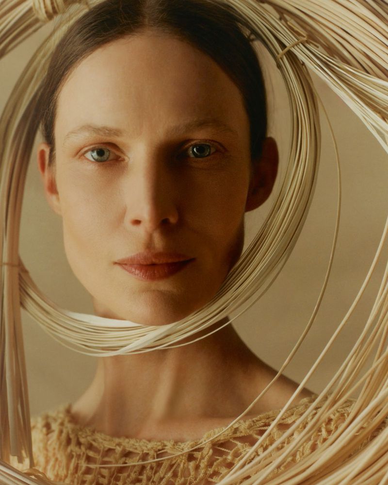 Close-up portrait of a fair-skinned adult woman framed by woven straw or wire. She wears a knitted top against a neutral backdrop.