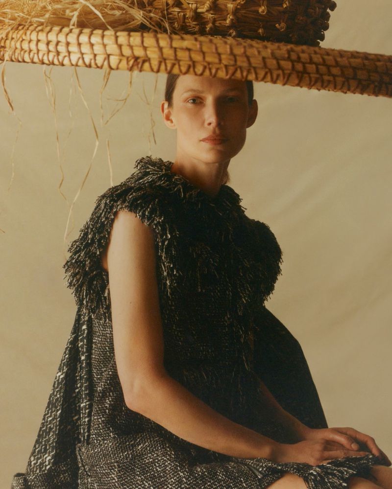 Model wearing a textured dark dress and wide straw hat against a warm neutral studio background