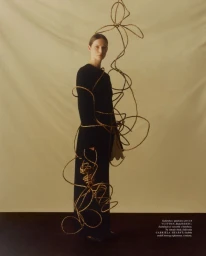 Model in a black dress standing against a neutral backdrop intertwined with looping wicker sculpture elements for Vogue Polska