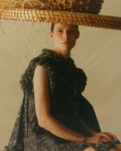 Model wearing a textured dark dress and wide straw hat against a warm neutral studio background