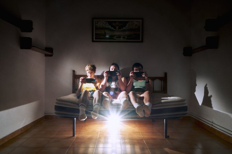 Three young adults are sitting on a bed in a dark room playing video games on handheld consoles, illuminated by a bright light source.