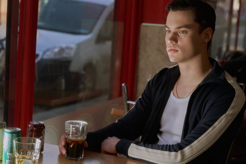 A young man sits inside a restaurant with drinks on the table, his gaze directed toward the window, with a blurred vehicle outside.