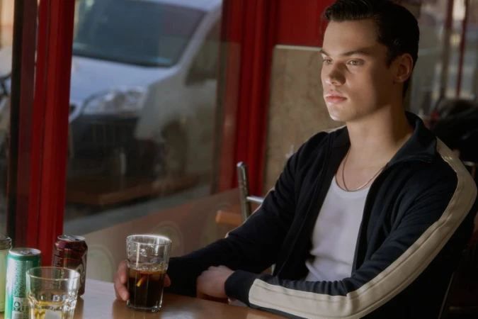 A young man sits inside a restaurant with drinks on the table, his gaze directed toward the window, with a blurred vehicle outside.