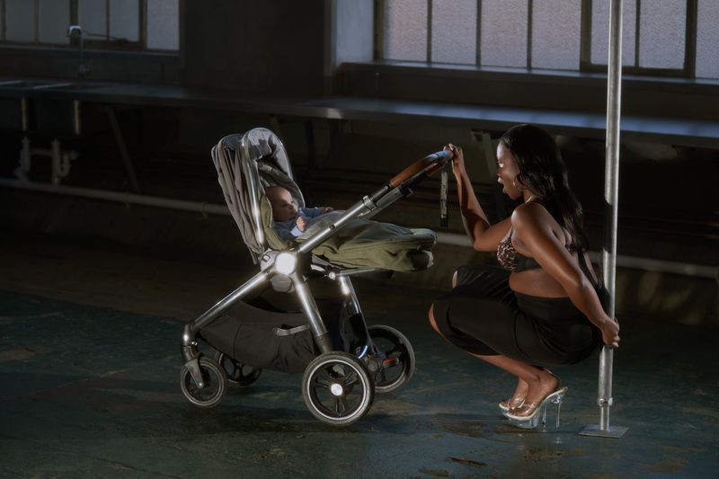 An adult female poses by a pole next to a baby in a stroller inside an empty industrial building with concrete floors.