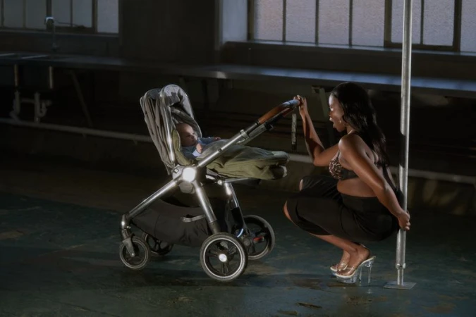 An adult female poses by a pole next to a baby in a stroller inside an empty industrial building with concrete floors.