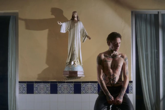 Shirtless man with tattoos sitting in front of statue, with blue and white tiled wall and bright lighting indoors.