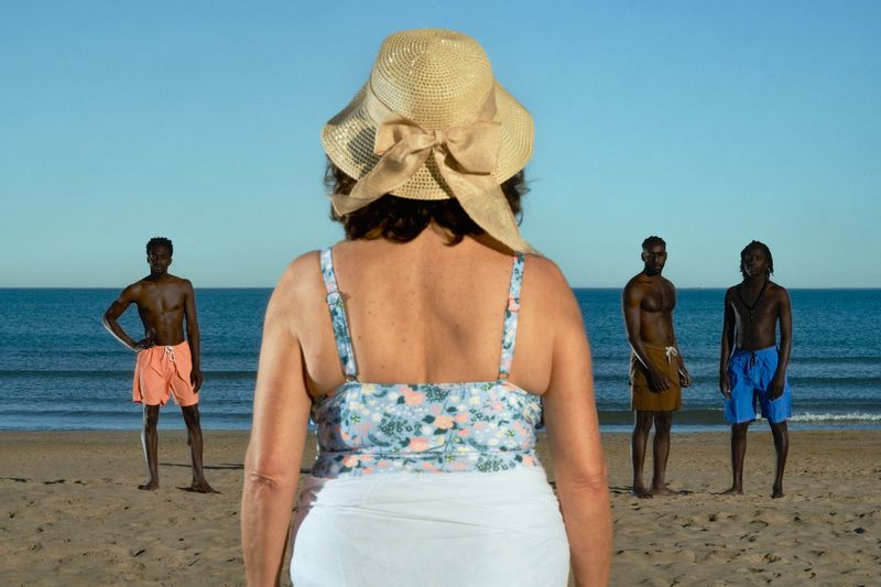 Adult woman in sunhat faces three black men on beach in swimwear on sunny day, clear sky and ocean in background, eye-level shot.