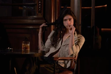 Model Lily Nova seated at a table in a bar playing with her long dark hair for Coeval Magazine STIGMATA