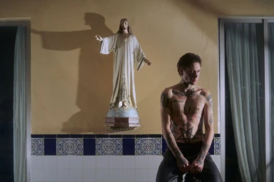Shirtless tattooed man seated in front of a yellow wall with a religious statue and patterned tile wainscoting.