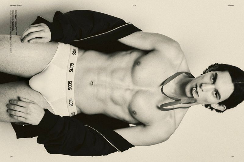 Topless man poses in underwear, wearing a jacket and ribbon, against a light backdrop in a studio setting for a fashion editorial.