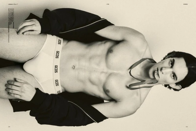 Topless man poses in underwear, wearing a jacket and ribbon, against a light backdrop in a studio setting for a fashion editorial.