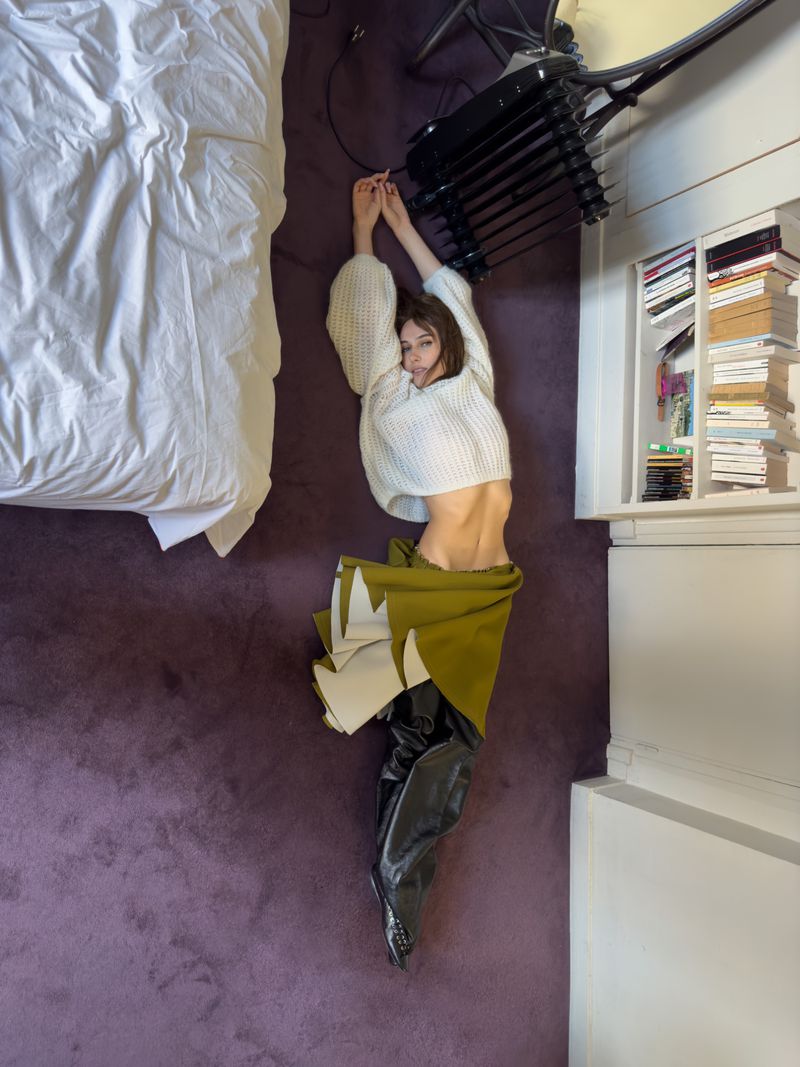Woman in white sweater, green skirt, and black boots lies on a purple rug near a white bed and stacked books in a neutral room.