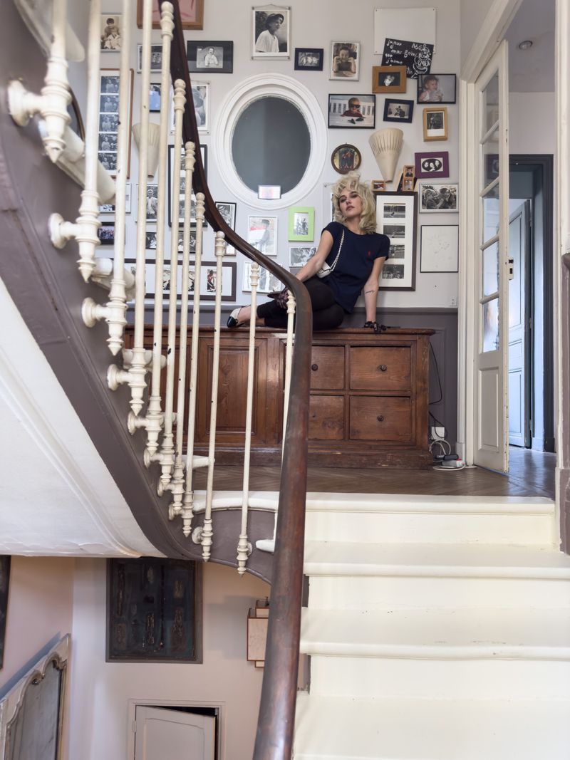 Woman with short blonde hair poses on a brown wooden dresser in a hallway with pictures on the wall and a staircase in the foreground.