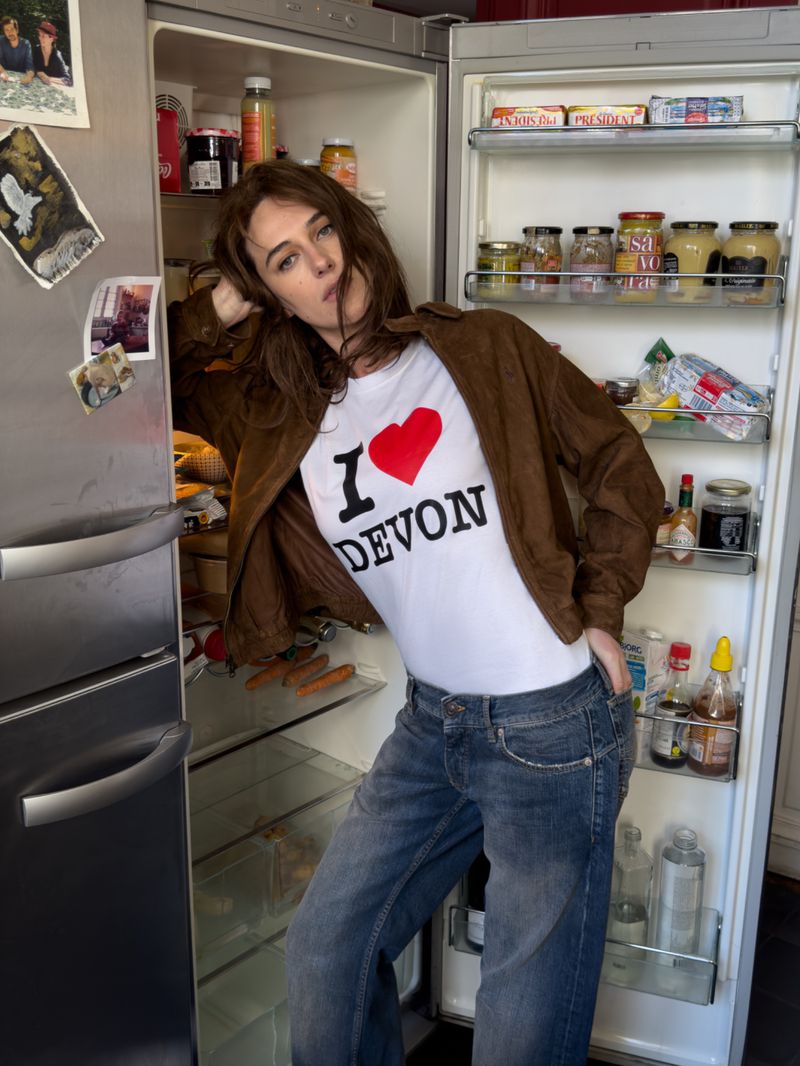 Adult woman in a brown jacket and jeans poses next to an open refrigerator filled with condiments, sauces, and jars inside an indoor space.