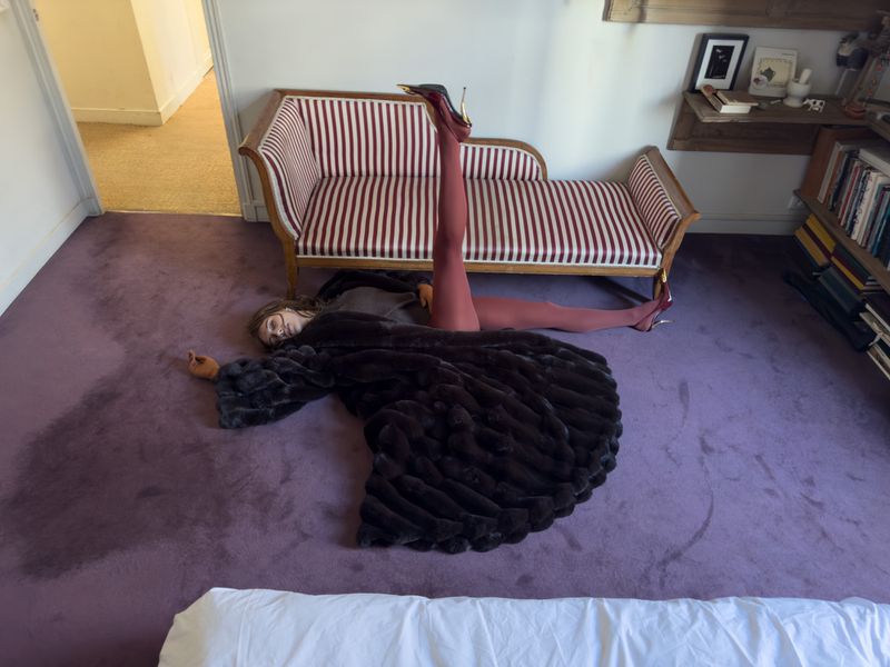 A woman in a fur coat and red tights is sprawled on the floor in a room with a striped settee, bookshelf, and lavender carpet.