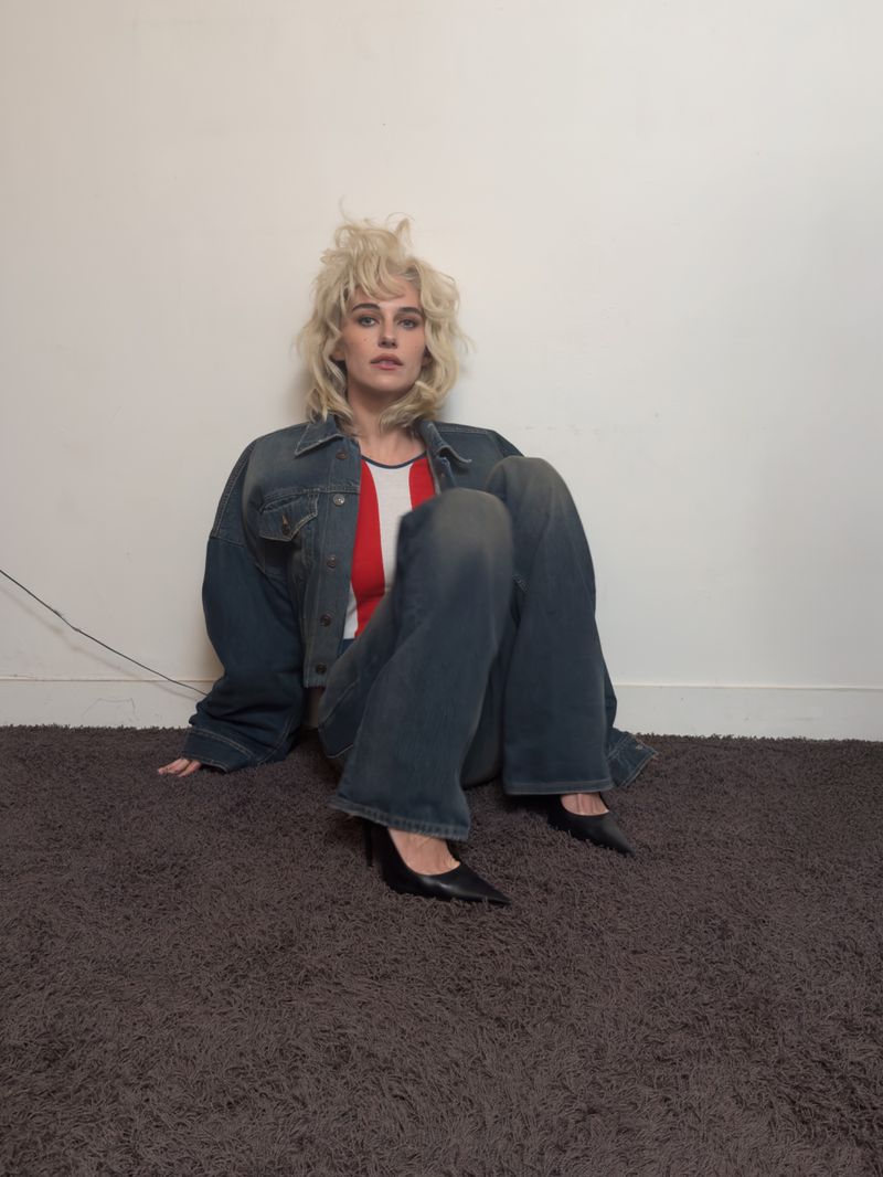 A young woman with blond hair sits on a gray carpet, wearing a denim jacket, striped shirt, blue jeans, and black heels, against a white wall.
