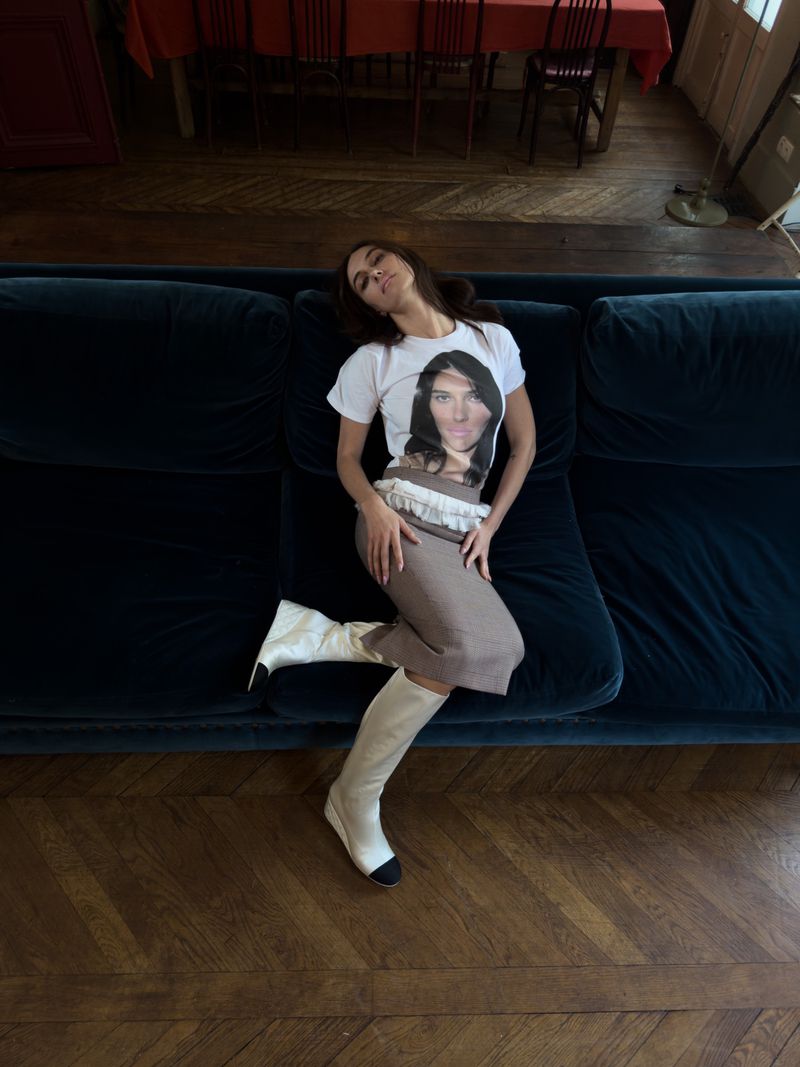Woman reclining on a blue velvet sofa wearing a graphic tee, pencil skirt, and knee-high boots in a room with hardwood floors.