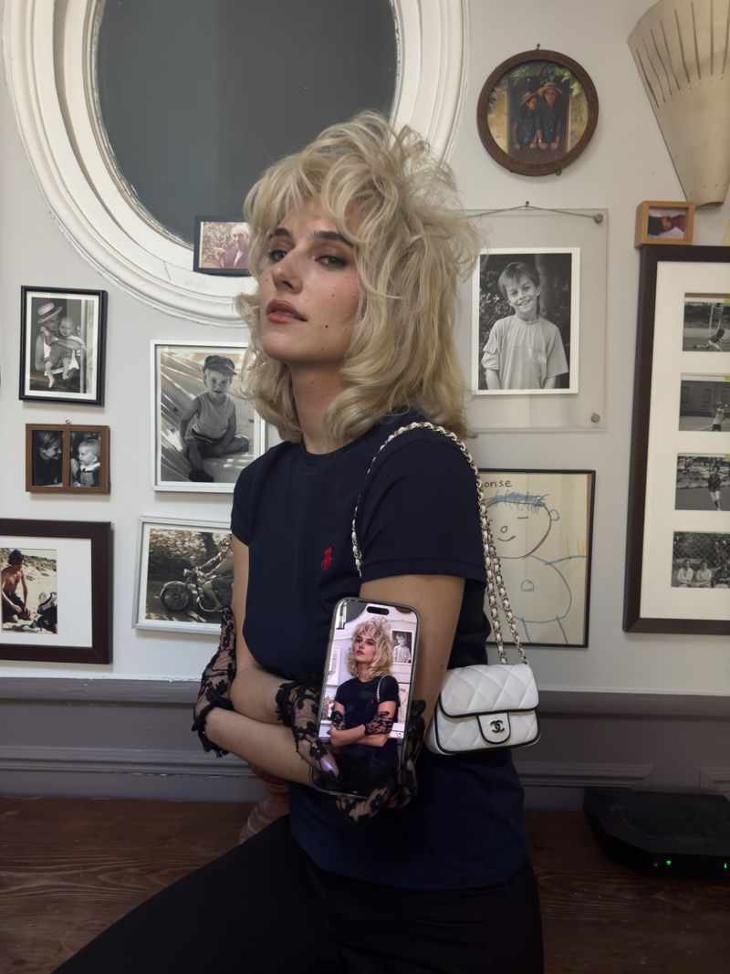 Woman with blonde hair wearing a black shirt and lace gloves holding a smartphone in front of a wall with framed photos.