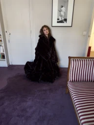 Devon Lee Carlson wearing a voluminous dark fur coat in a room with a striped sofa and framed artwork.
