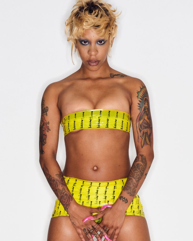 Full shot of a woman with blonde hair wearing a yellow tube top and skirt, posing in a photo studio against a white backdrop.