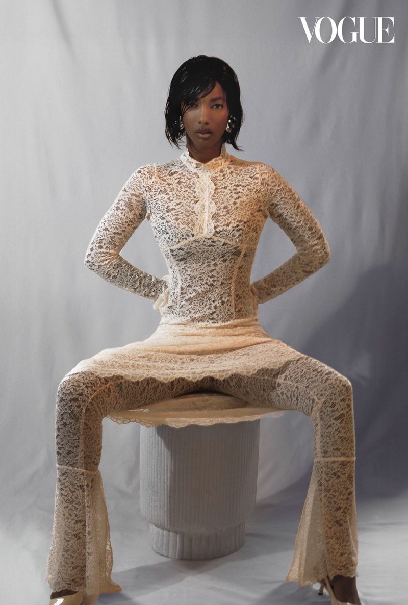 A stylish woman in a lace bodysuit sits confidently, showcasing fashion against a muted backdrop, creating a Vogue-worthy editorial image.
