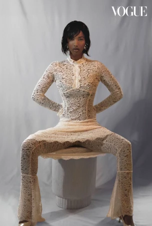 A stylish woman in a lace bodysuit sits confidently, showcasing fashion against a muted backdrop, creating a Vogue-worthy editorial image.
