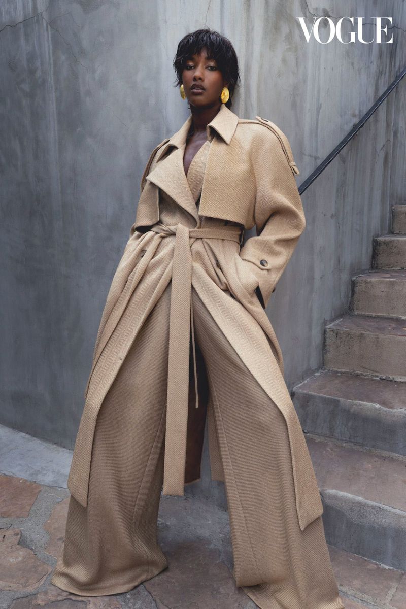 A black female model poses on stone steps wearing a beige belted trench coat, wide-leg pants, and yellow earrings for a fashion editorial.