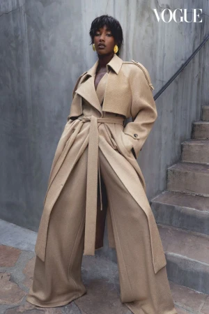 A black female model poses on stone steps wearing a beige belted trench coat, wide-leg pants, and yellow earrings for a fashion editorial.