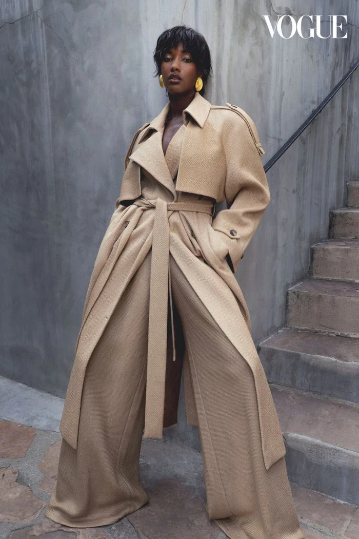 Model wearing a tan trench coat and wide-leg trousers standing against a textured concrete wall beside stone stairs.