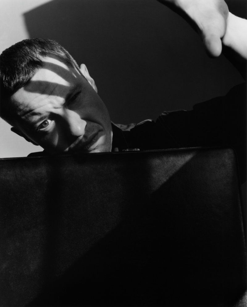 Monochrome portrait of a man holding a briefcase, casting shadows on his face. The photo features strong lighting and contrasts.